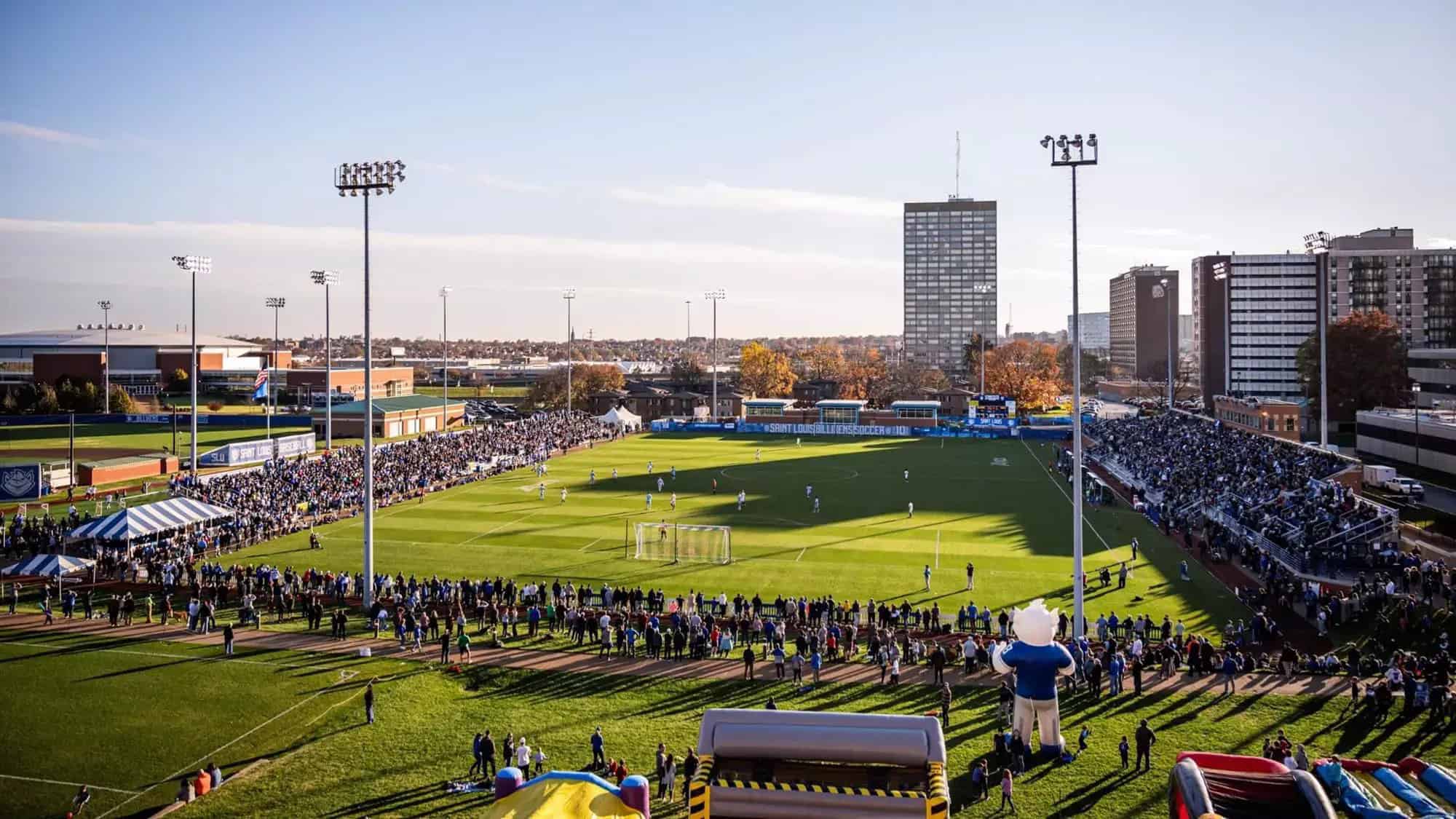 NCAA college soccer game at Saint Louis University with full stands, campus buildings, and players on the field